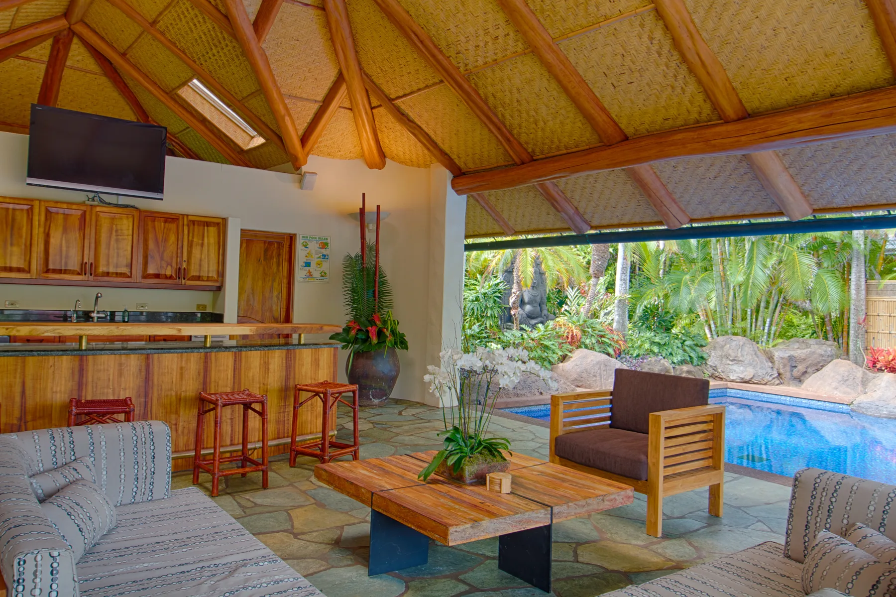 Una sala y cocina al aire libre dan a una piscina rodeada de vegetación tropical.