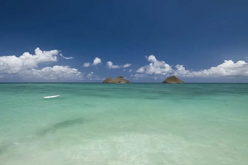 Un mar turquesa se extiende hacia dos pequeños islotes rocosos bajo un cielo azul con nubes.