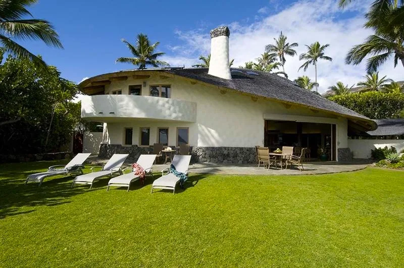 Una casa tropical con techo curvo da a un césped con asientos al aire libre.