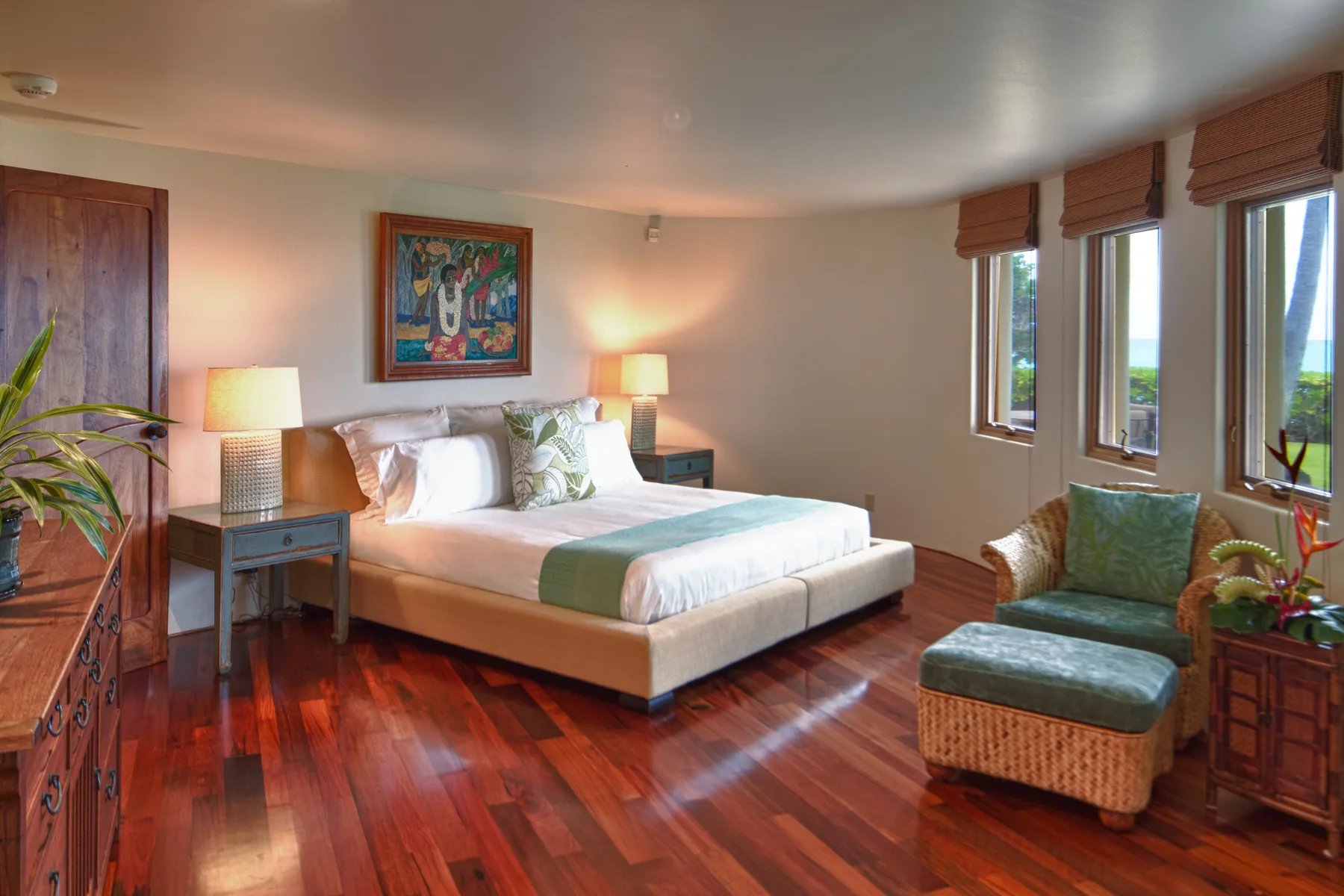 Un dormitorio con una cama, mesitas de noche y un sillón verde junto a las ventanas.