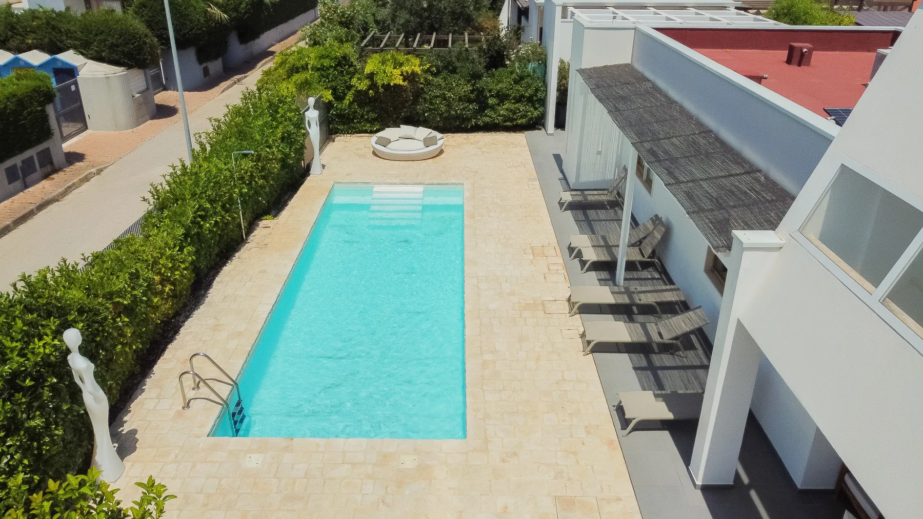 Una piscina al aire libre junto a un edificio, con suelo pavimentado y vegetación.