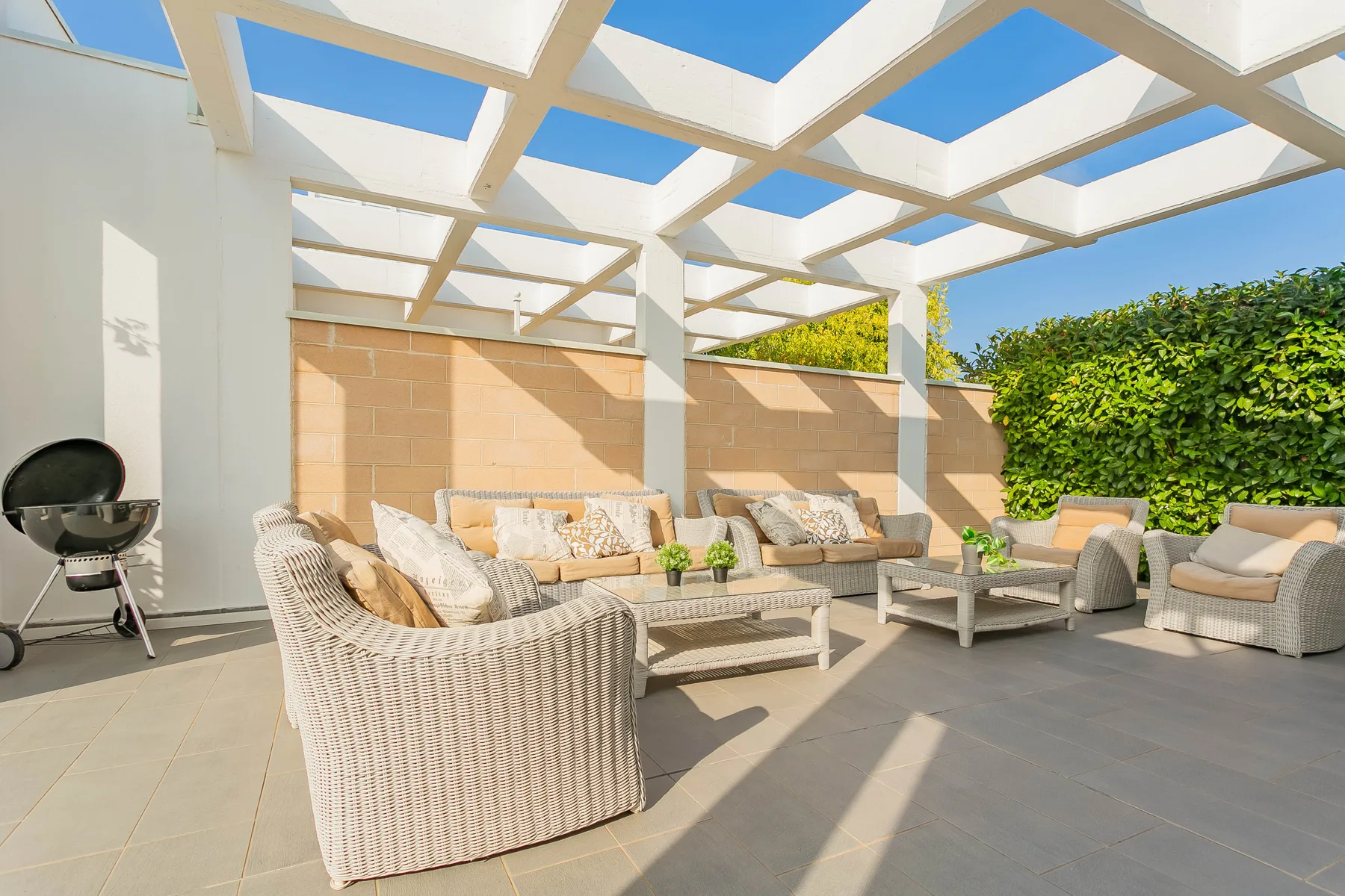 Una zona de estar exterior soleada bajo una pérgola blanca, con muebles acolchados.