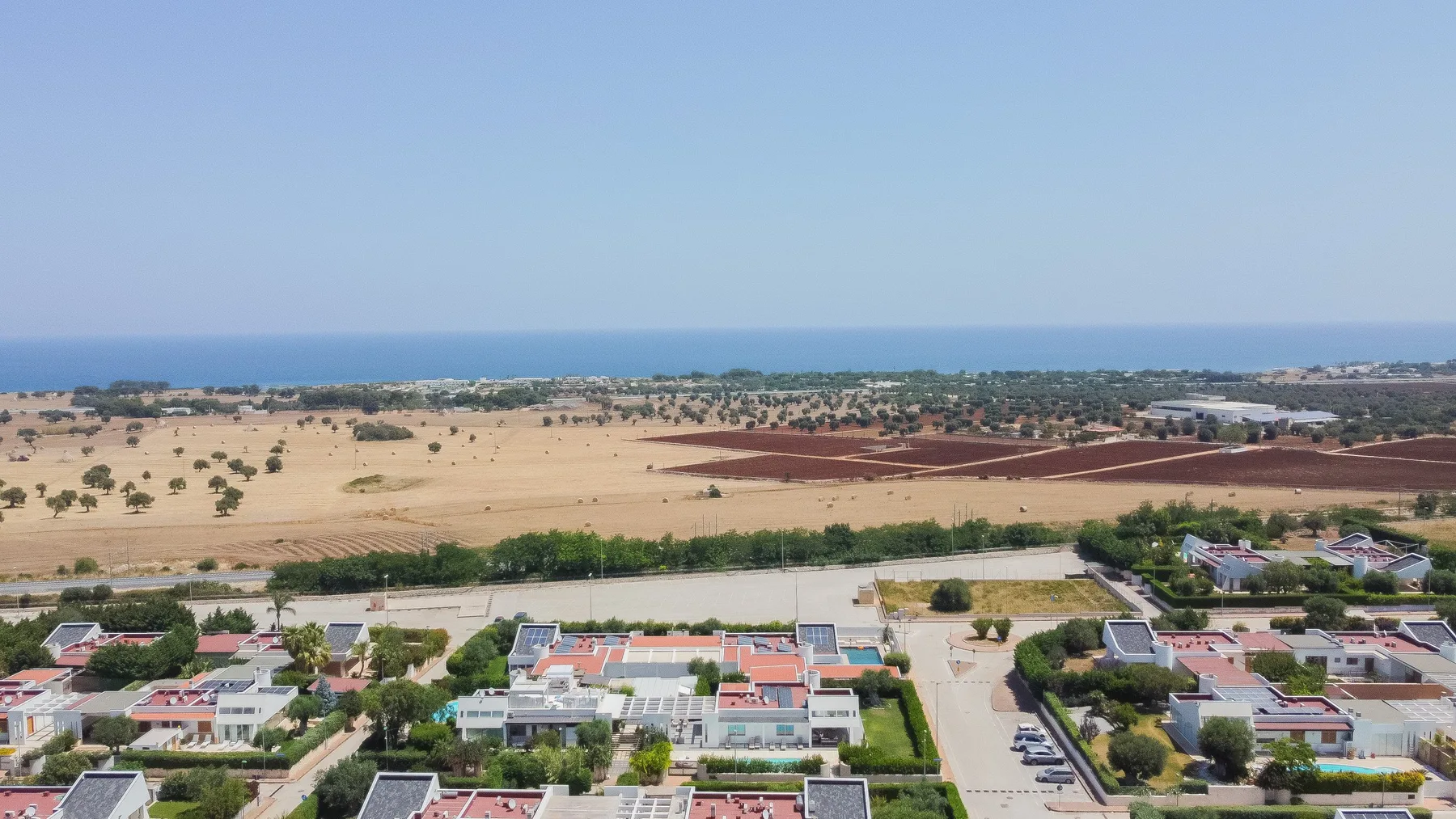 Vista aérea de un barrio residencial junto a campos abiertos, con el mar en el horizonte.