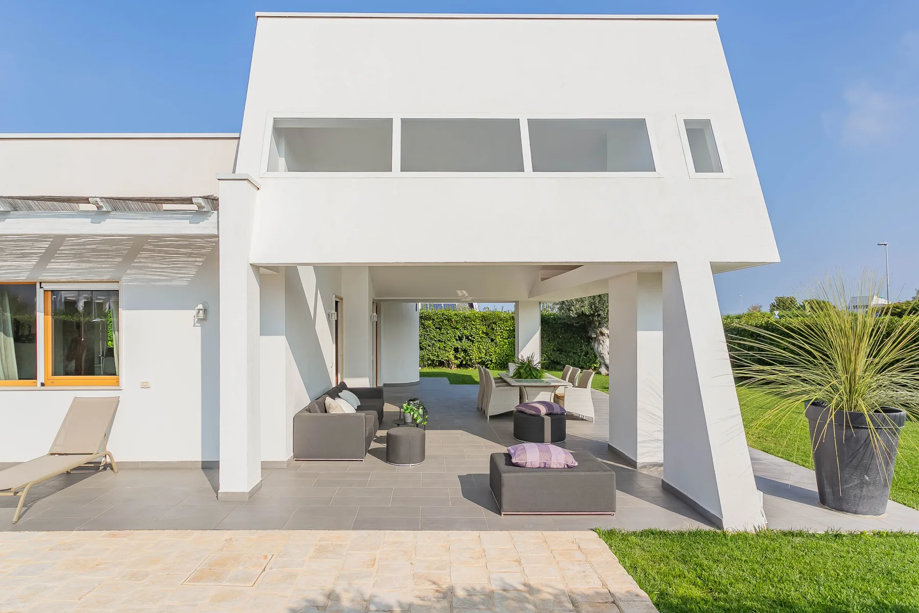 Una casa blanca moderna se abre a un patio cubierto con asientos al aire libre y un jardín.