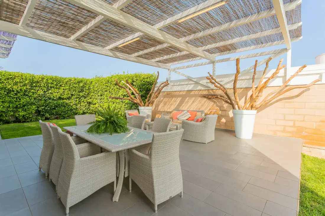 Un patio exterior sombreado tiene una mesa de comedor, asientos y plantas en macetas.