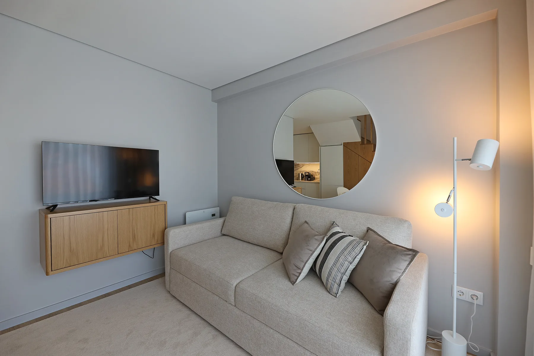 A living room with a beige sofa, wall-mounted television, and round mirror.