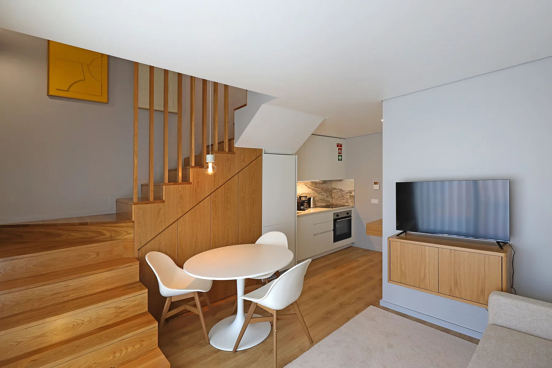 A compact living area has a dining table, wooden staircase, and television.