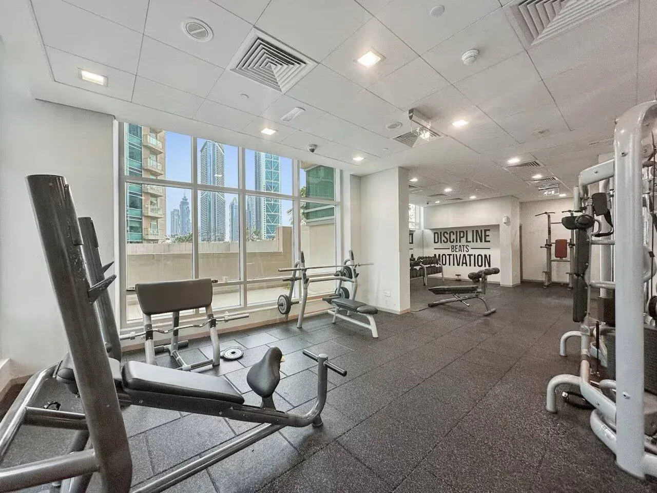 Un gimnasio con equipo de ejercicio y grandes ventanas con vistas a edificios cercanos.