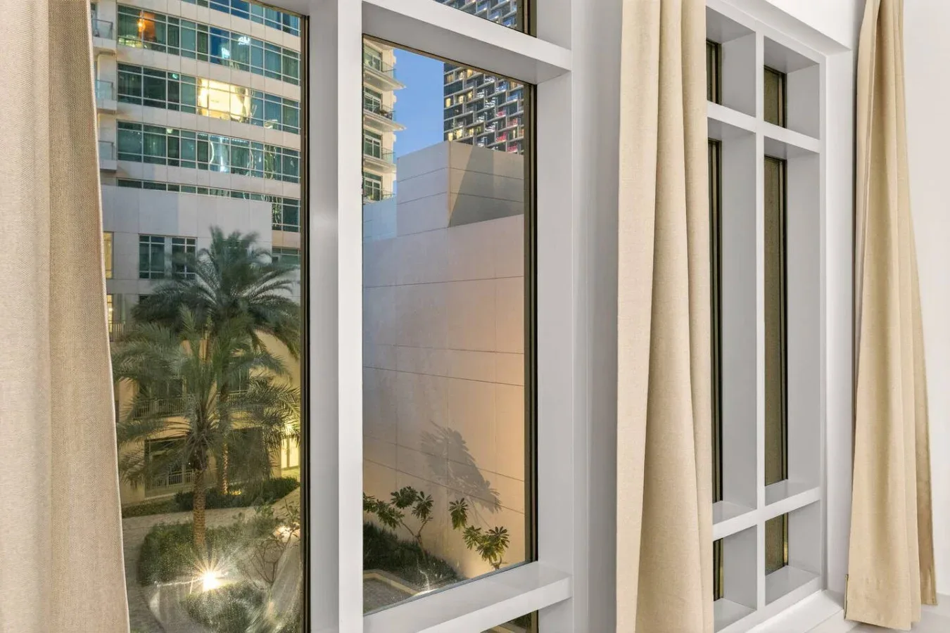 Vista de edificios cercanos y palmeras a través de una ventana con cortinas claras.