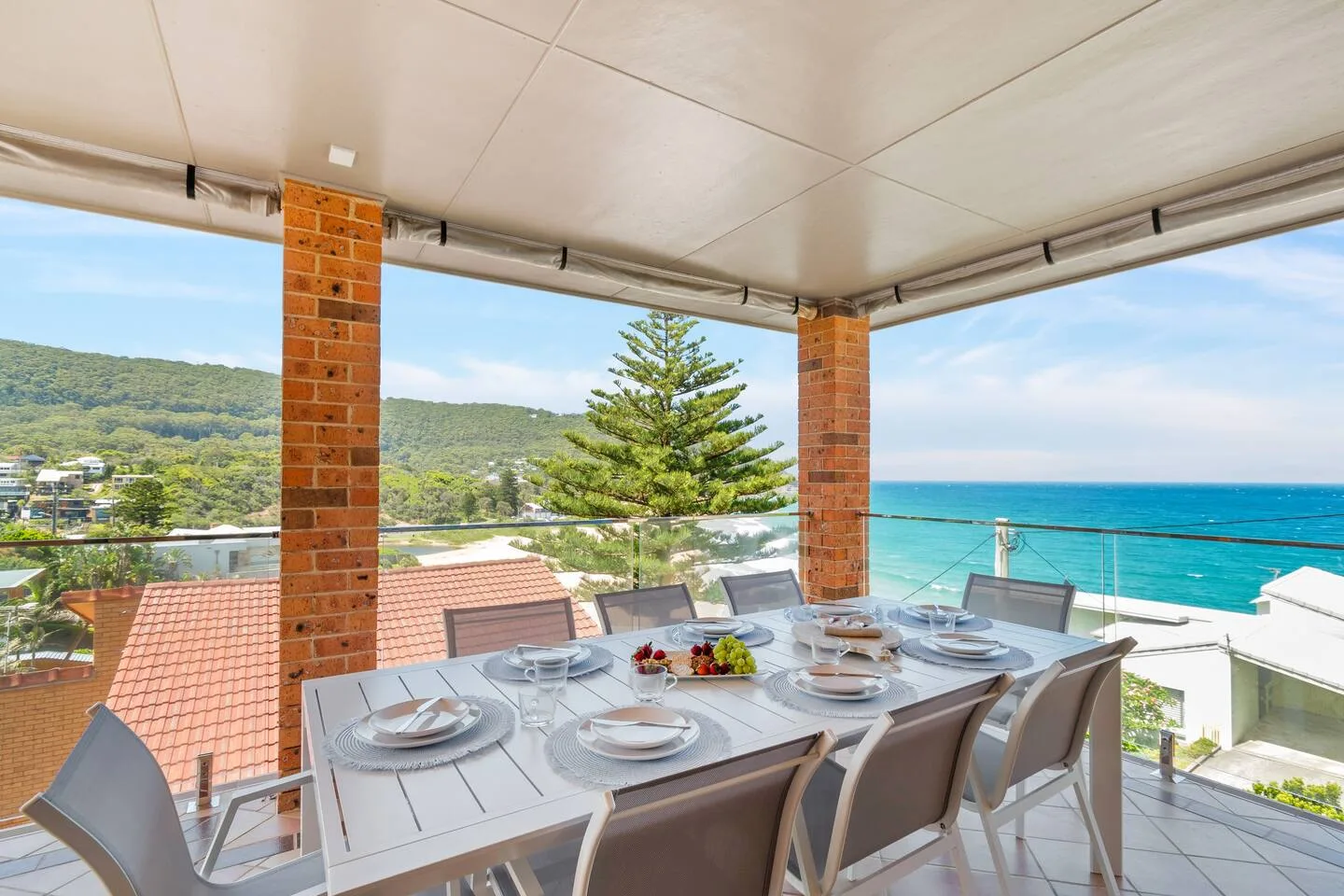 Una zona de comedor en una terraza cubierta tiene vistas al mar y a las colinas cercanas.