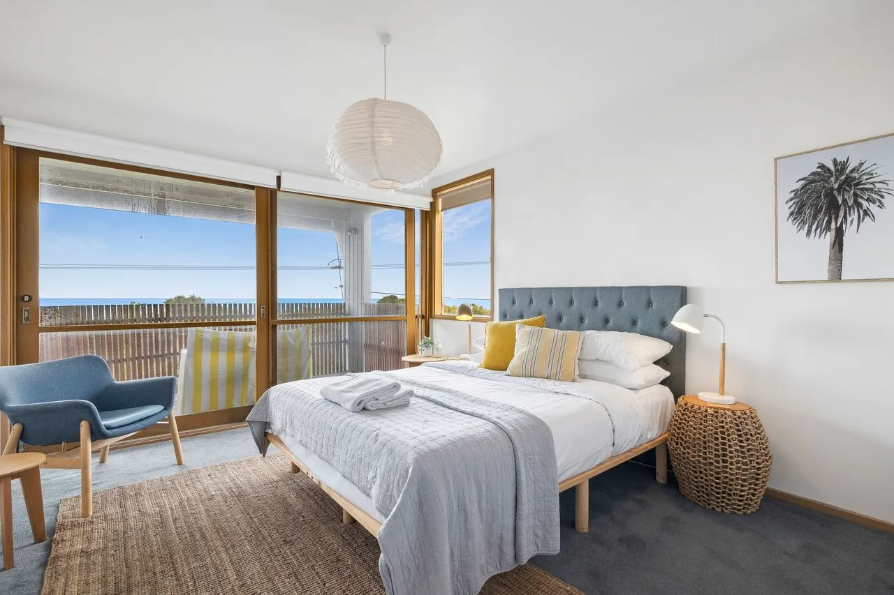 A bedroom with a bed beside wide windows overlooking the ocean.