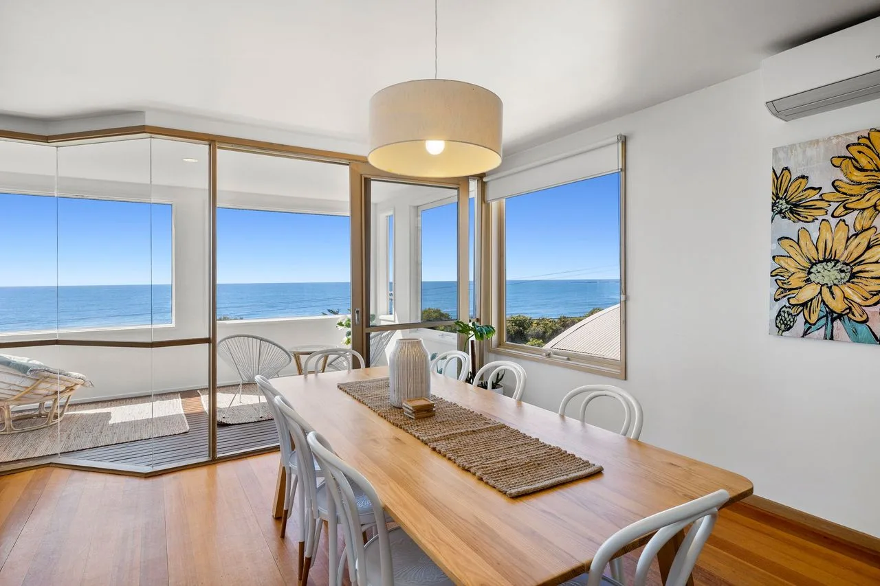 A dining area with a wooden table and chairs overlooks the ocean through large windows.