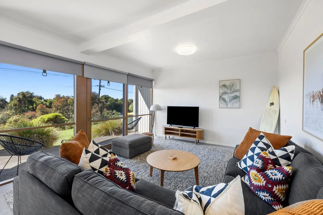 A living room with gray seating, a television, and large windows overlooking greenery.