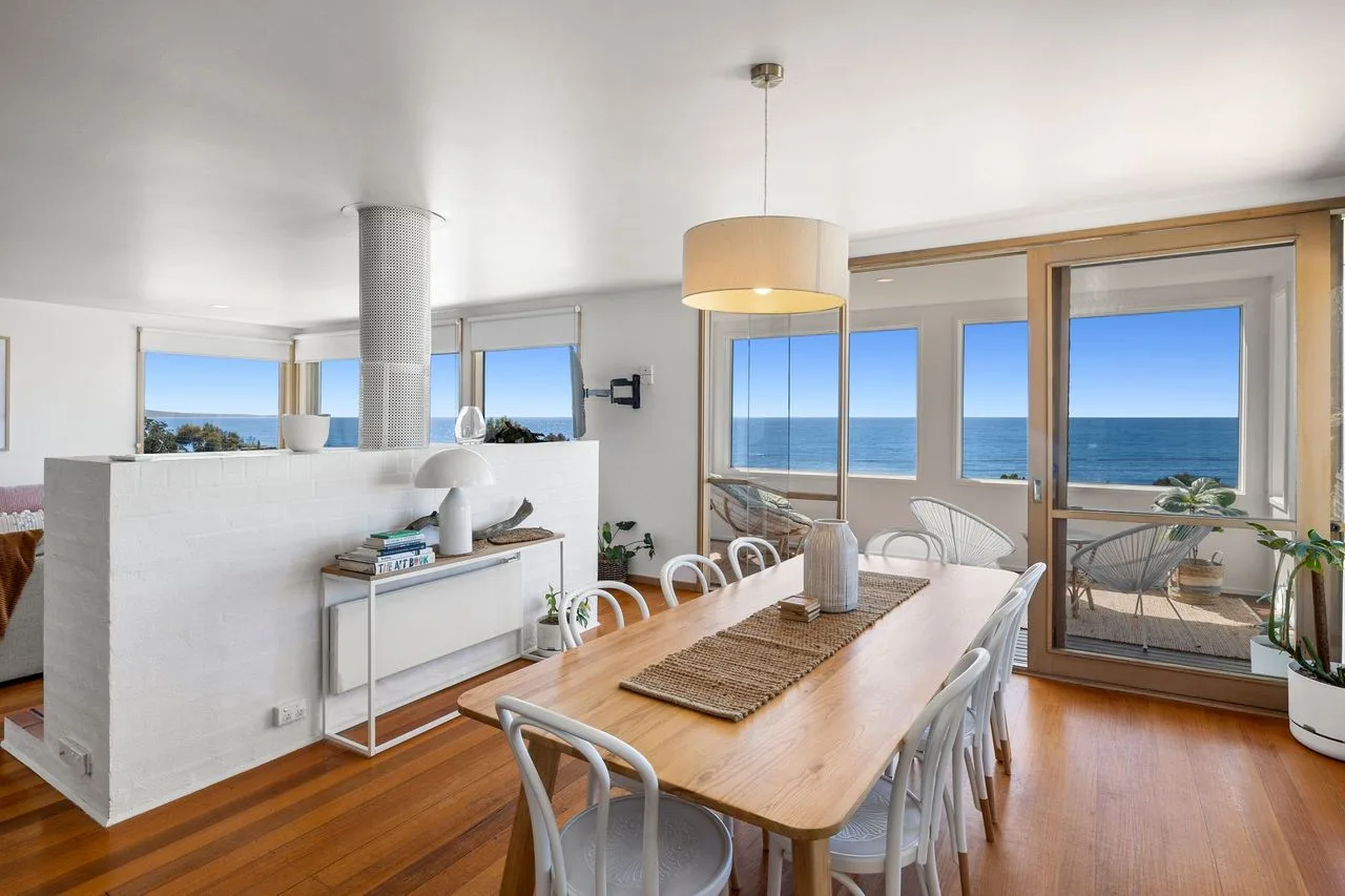A dining area with a long table beside large windows overlooking the ocean.