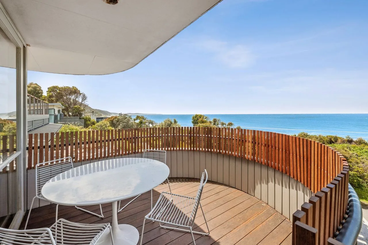 A curved outdoor deck with a table overlooks the ocean.