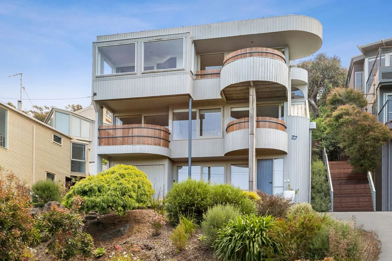 A modern multi-story building stands behind landscaped shrubs and trees.