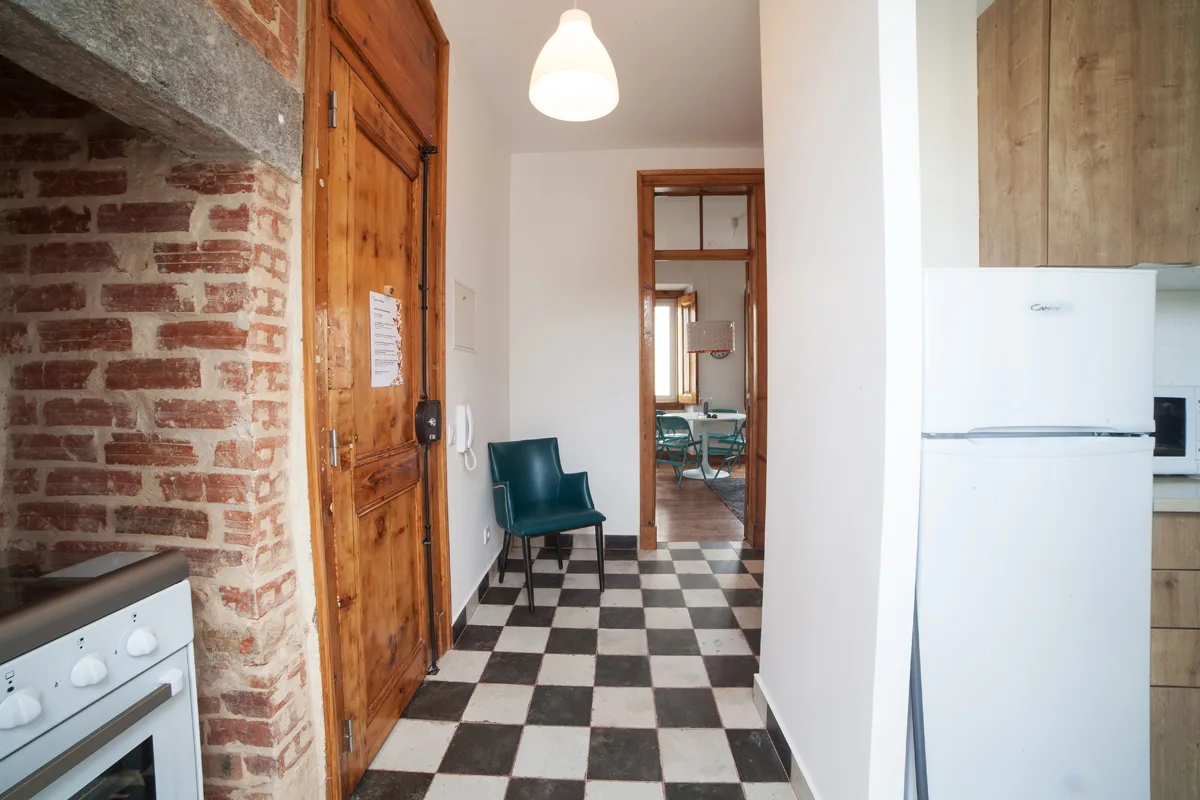 A narrow kitchen passage has a checkered floor, brick wall, and wooden door.