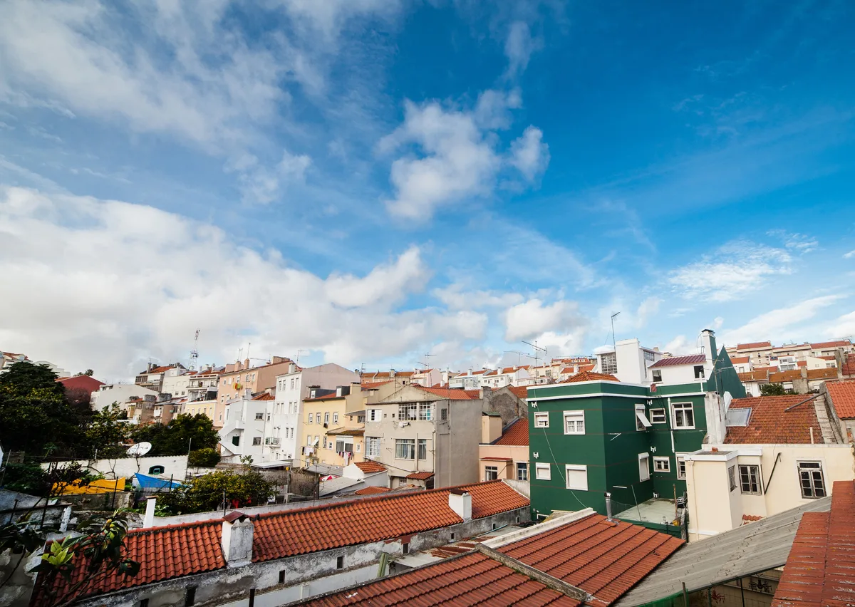 A cityscape of closely grouped buildings with red-tiled roofs beneath a blue, cloud-filled sky.