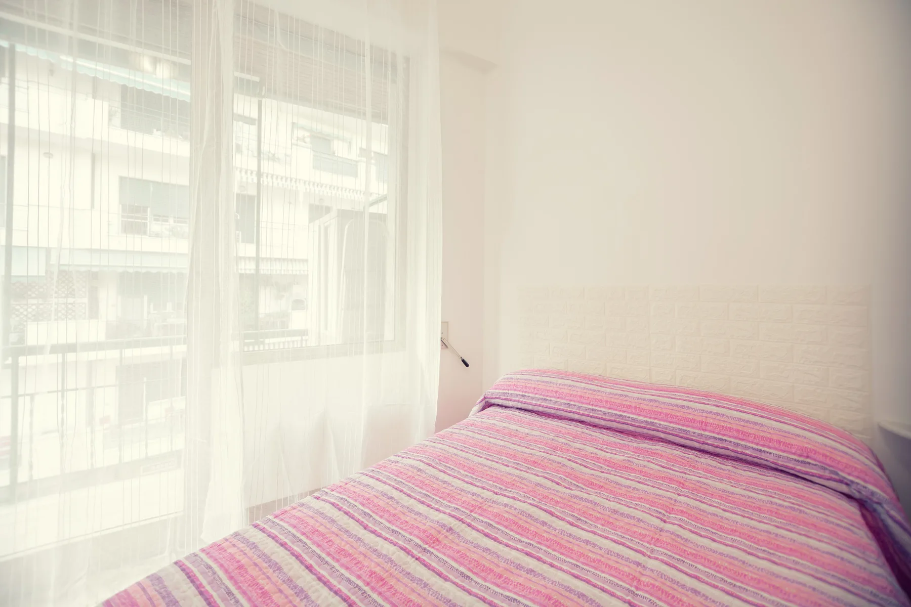 A bedroom with a bed covered in a pink striped bedspread beside sheer curtains.
