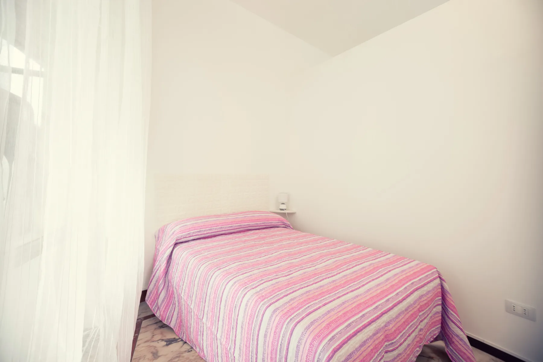 A bedroom with a bed covered in a pink striped bedspread beside a curtained window.