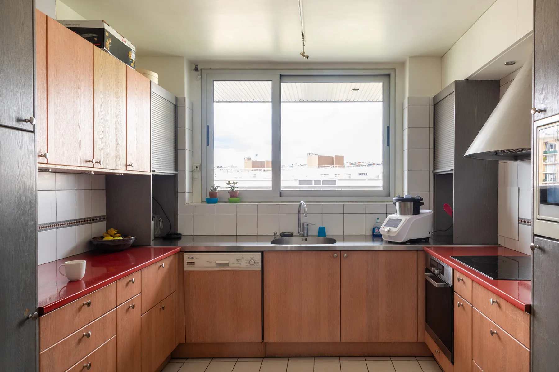 A kitchen with wooden cabinets and red countertops around a central window.