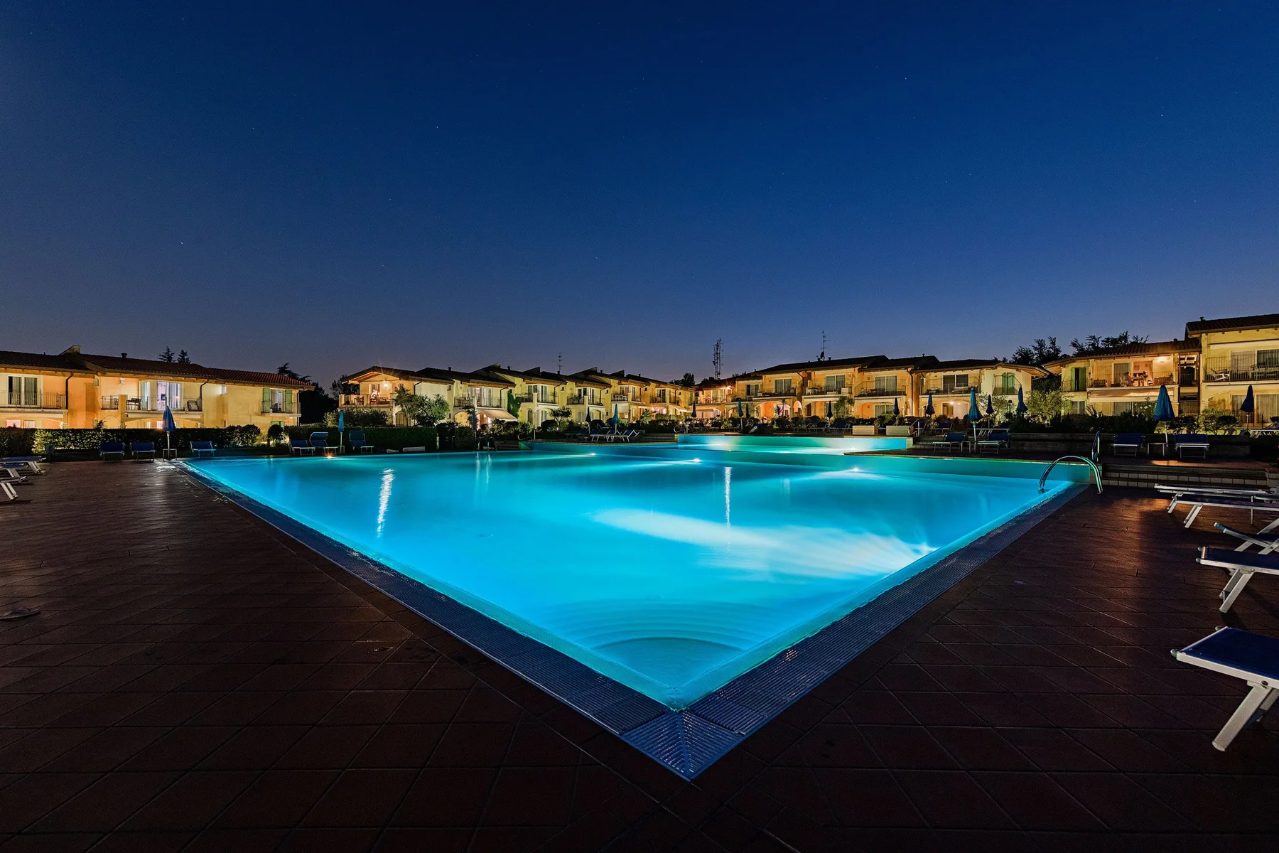 An illuminated swimming pool sits beside a deck with buildings in the background.
