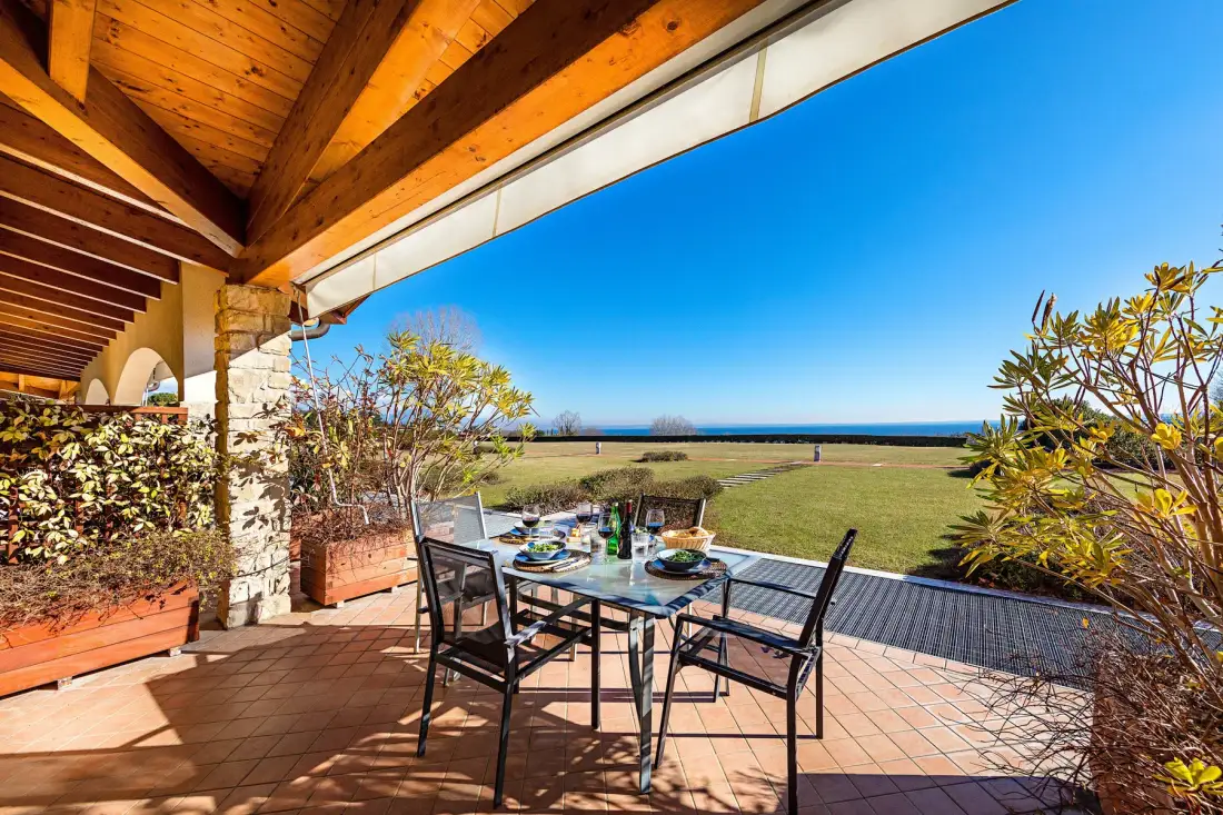 A covered outdoor dining area opens onto a grassy landscape and distant water.