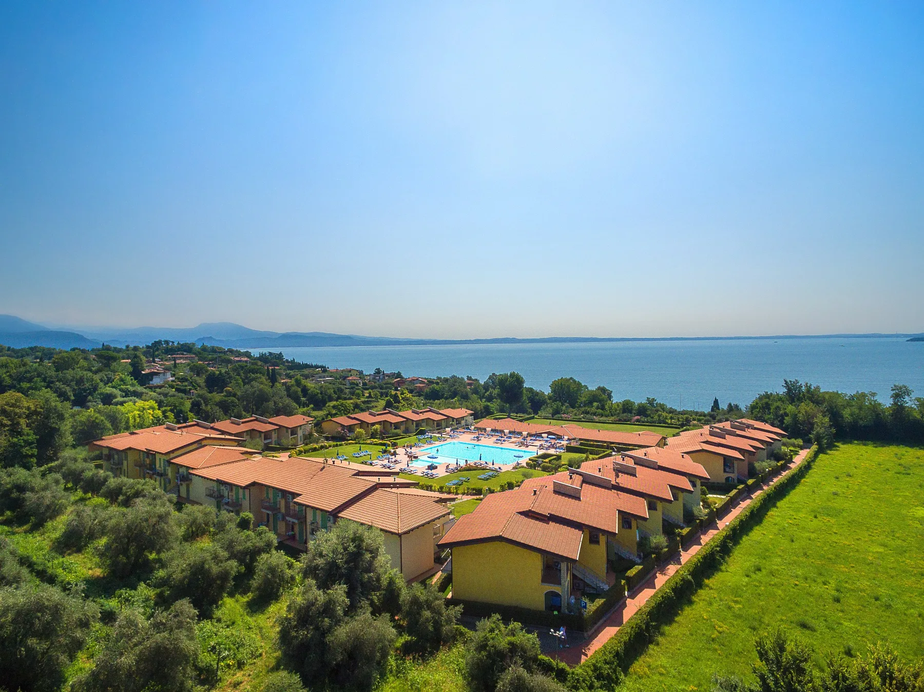 Aerial view of a resort with red-roofed buildings, a swimming pool, and the sea beyond.