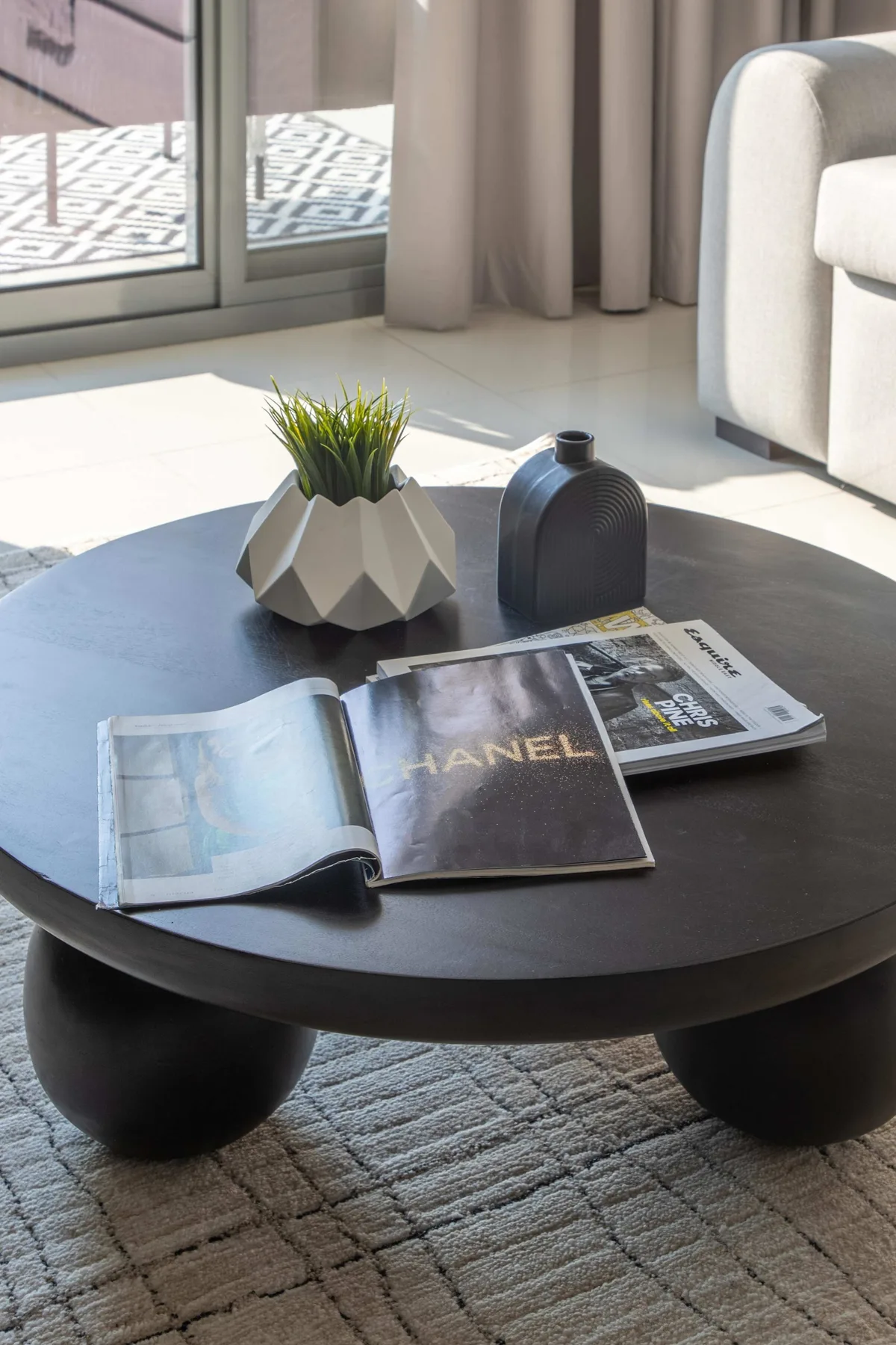 A round coffee table holds magazines and a small potted plant beside a sofa.