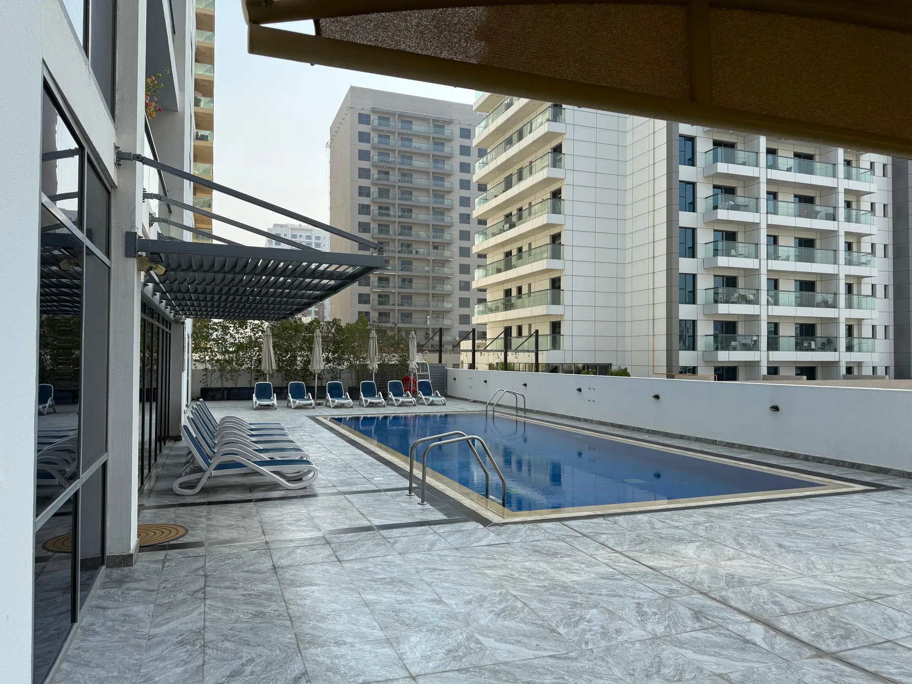 An outdoor swimming pool sits beside a paved deck and modern apartment buildings.