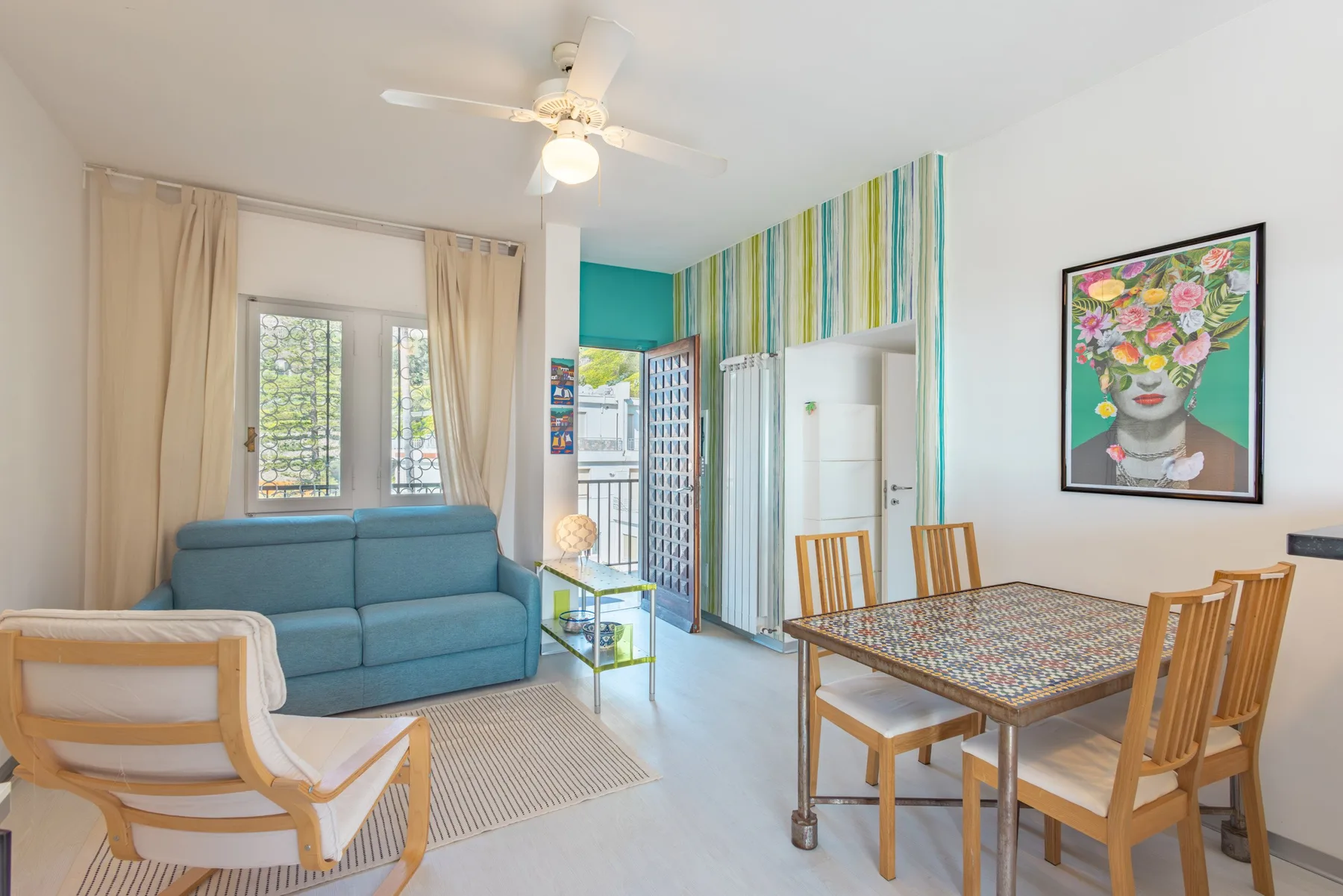 A bright living and dining area has blue seating, a patterned table, and colorful wall art.
