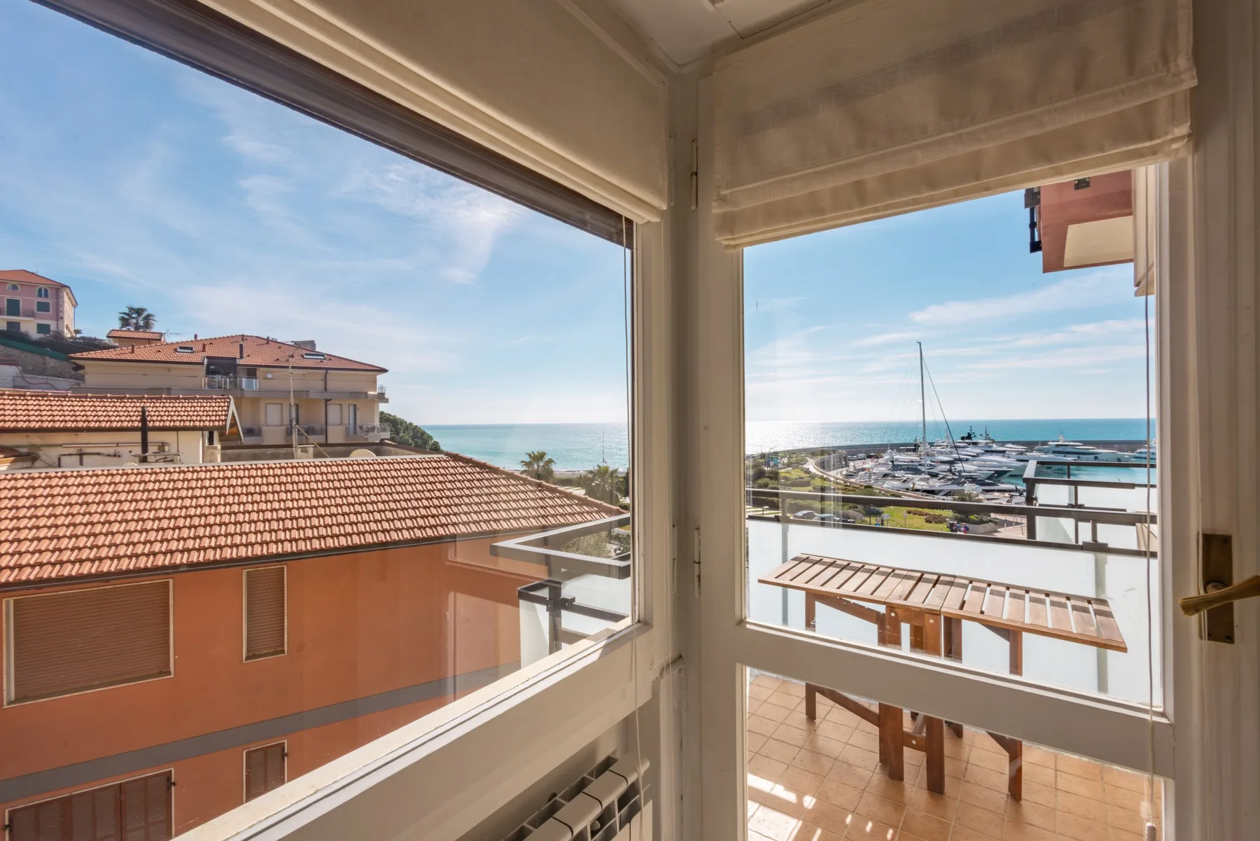 Una vista luminosa desde la ventana muestra tejados cercanos, el mar y un puerto deportivo.
