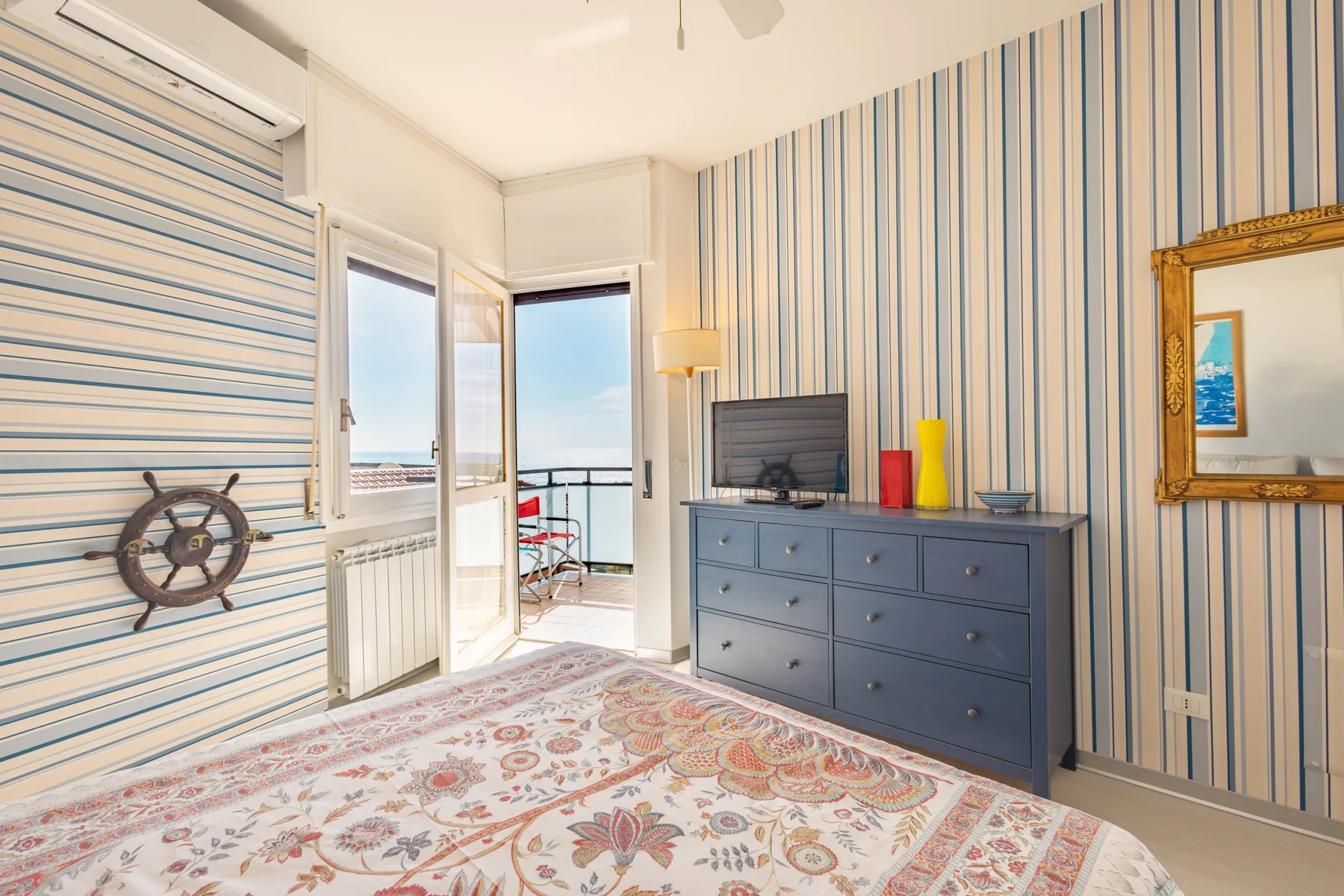 Un dormitorio luminoso con paredes a rayas y una puerta de balcón abierta al mar.