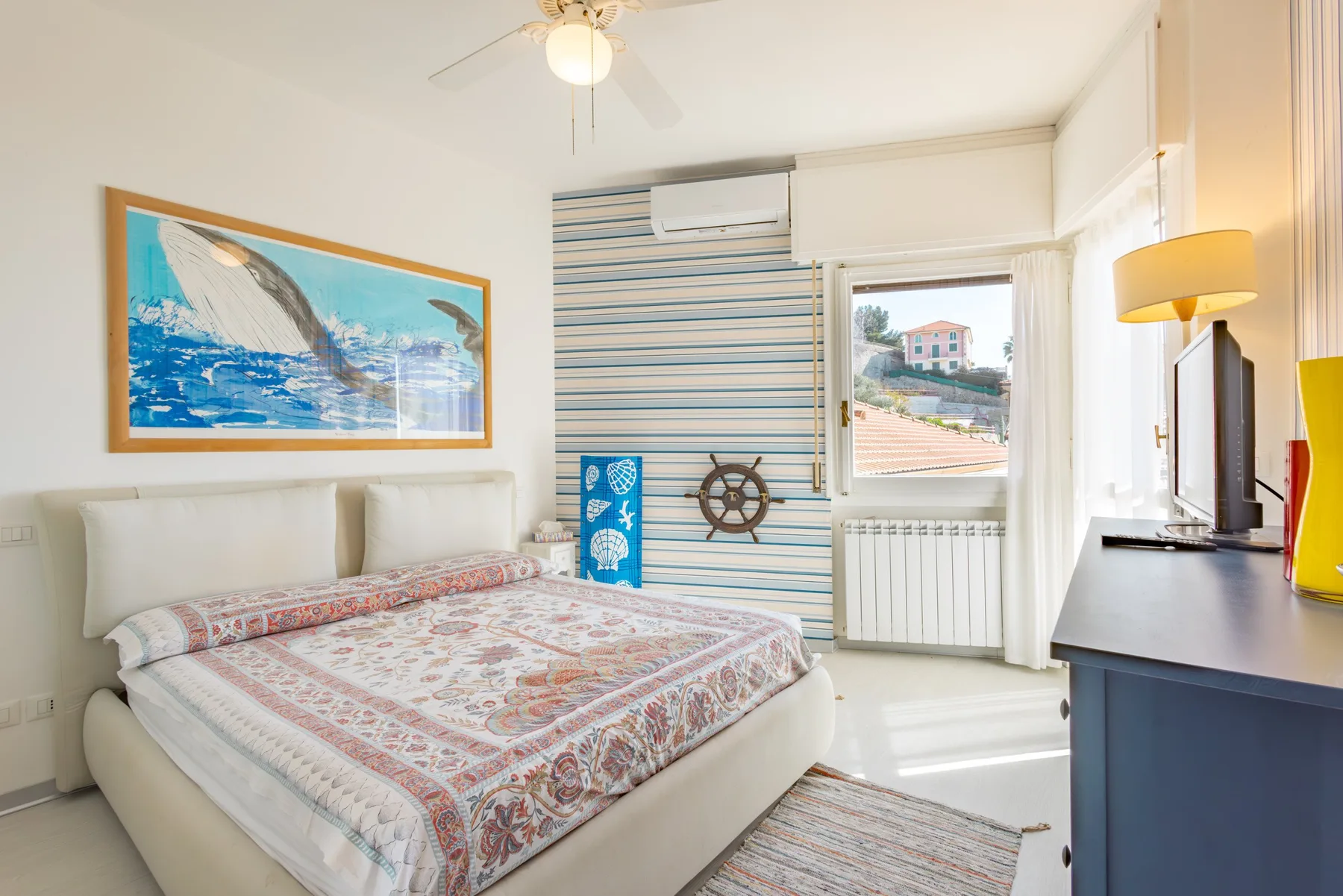 A bright bedroom with a patterned bedspread and a window overlooking nearby buildings.