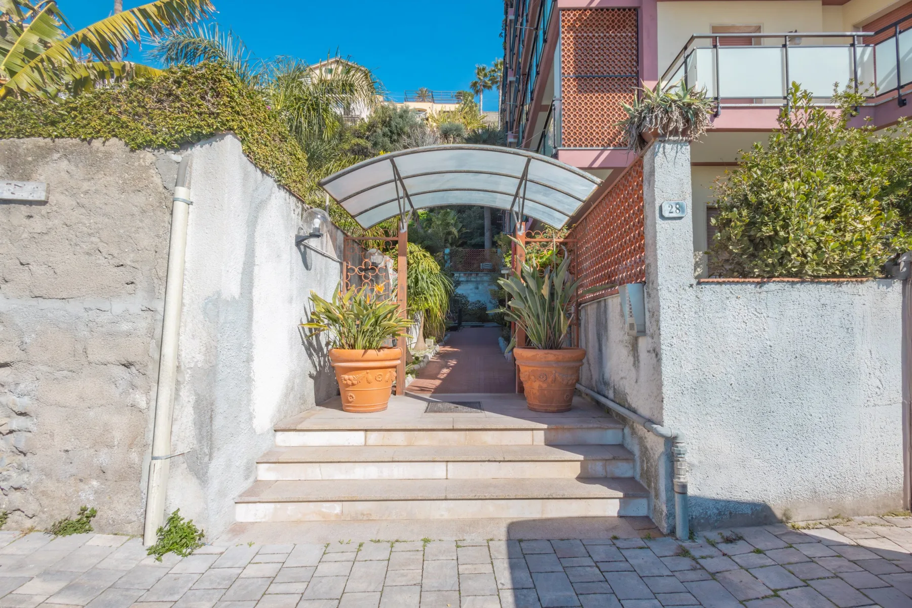 A paved entrance walkway leads up steps beneath a curved canopy, with potted plants.
