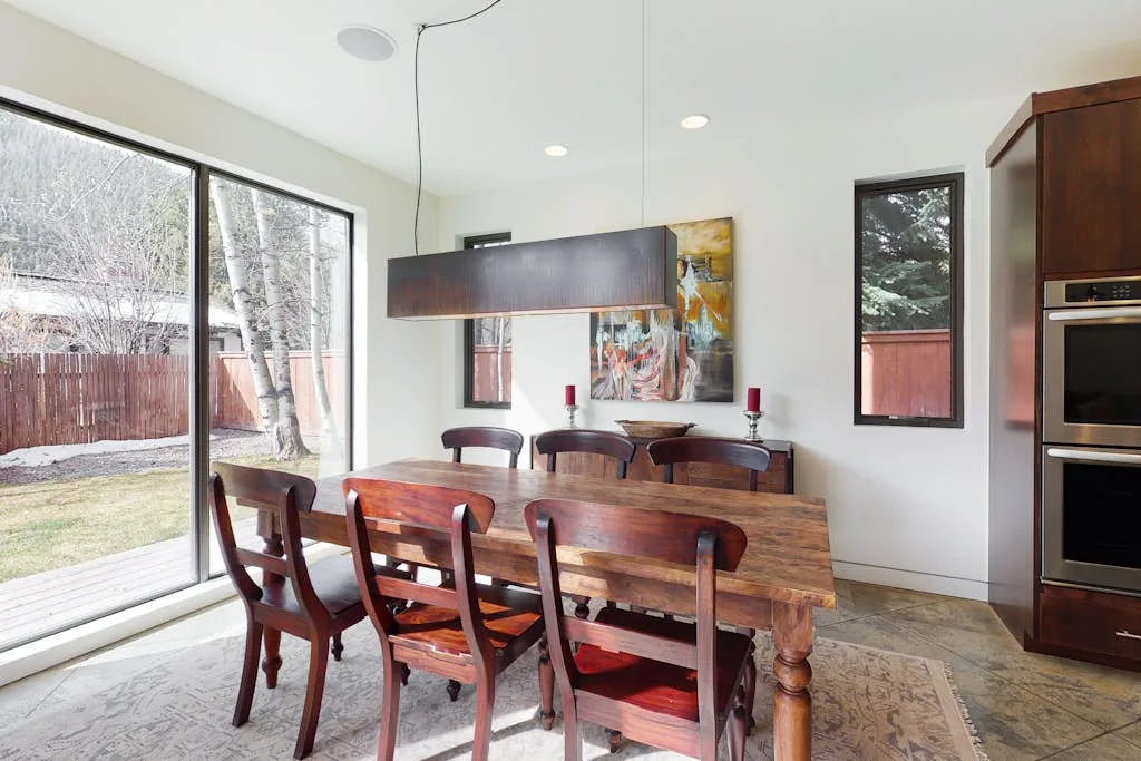 A dining area with a wooden table and chairs beside large windows.