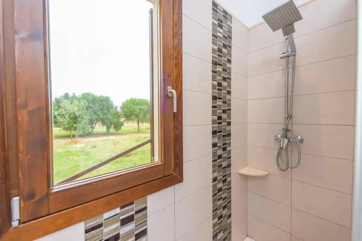 Una ducha con azulejos junto a una ventana de marco de madera con vistas a zonas verdes.
