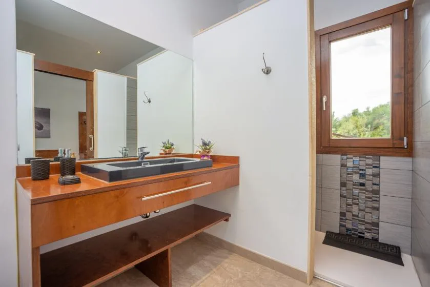 Un lavabo de baño con un espejo amplio está junto a una ventana y una zona de ducha con azulejos.