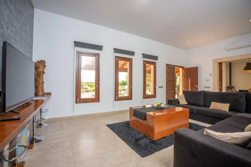 Una sala de estar con asientos oscuros, una mesa de centro de madera y ventanas altas.