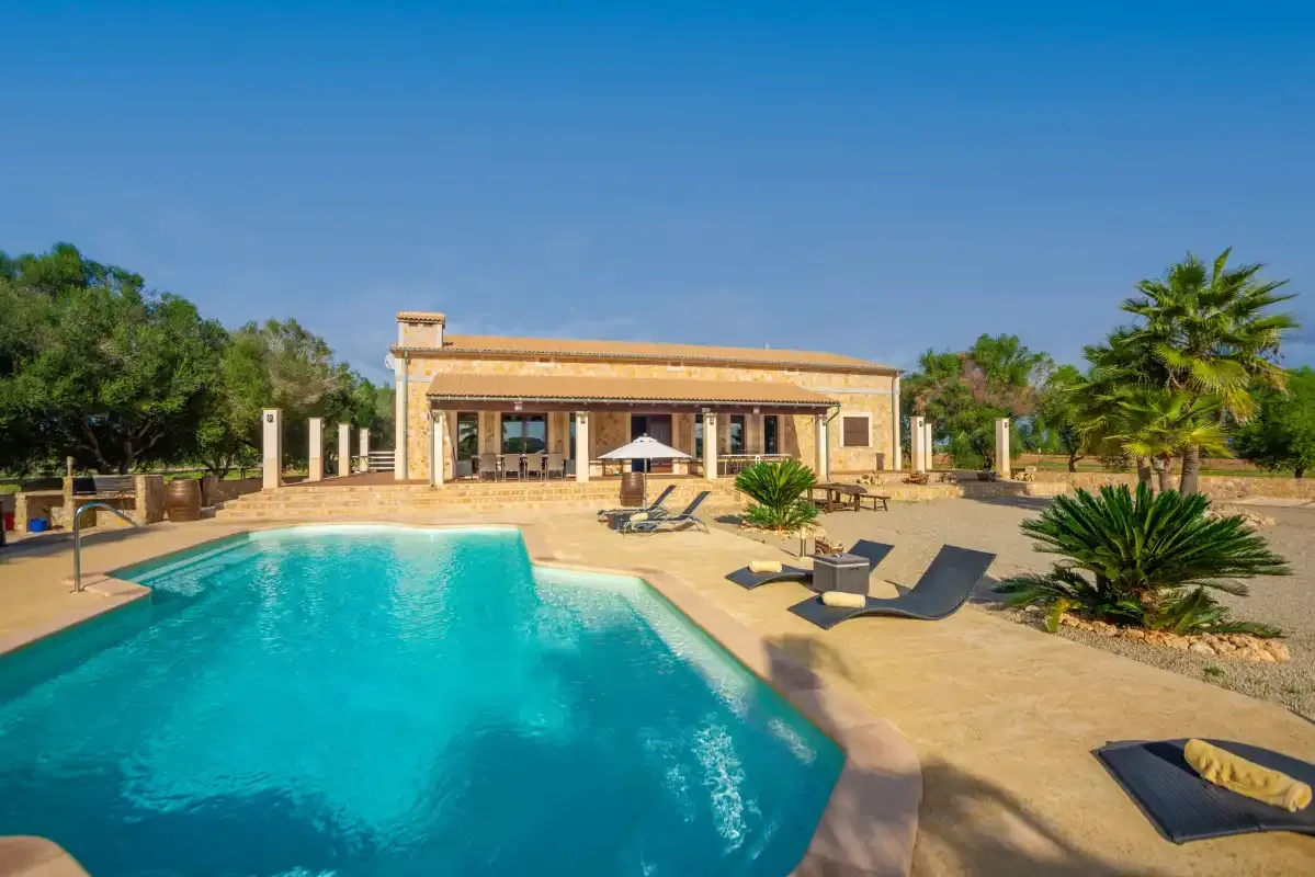Piscina al aire libre junto a una villa baja, con asientos y vegetación.