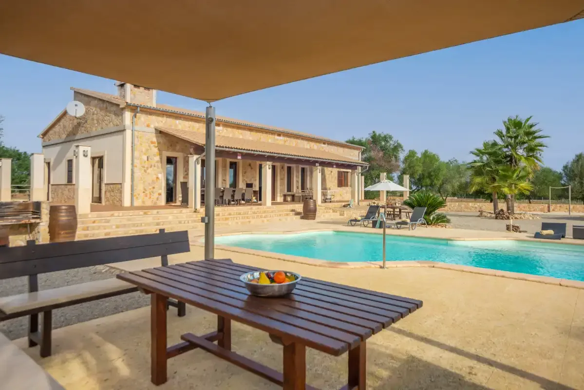 Un patio junto a la piscina con una mesa de comedor de madera y un edificio revestido de piedra.
