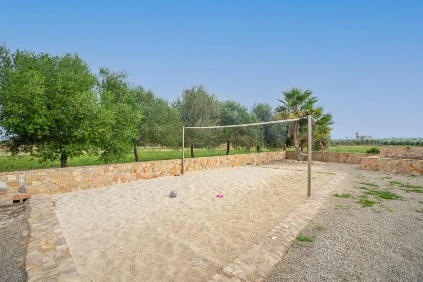 Una cancha exterior de arena con red está junto a un muro bajo de piedra y árboles.