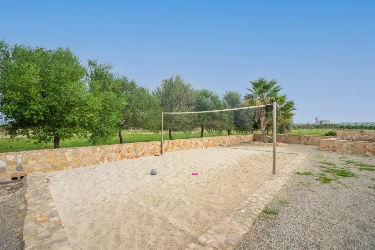 Una cancha exterior de arena con una red está junto a vegetación y un muro de piedra.