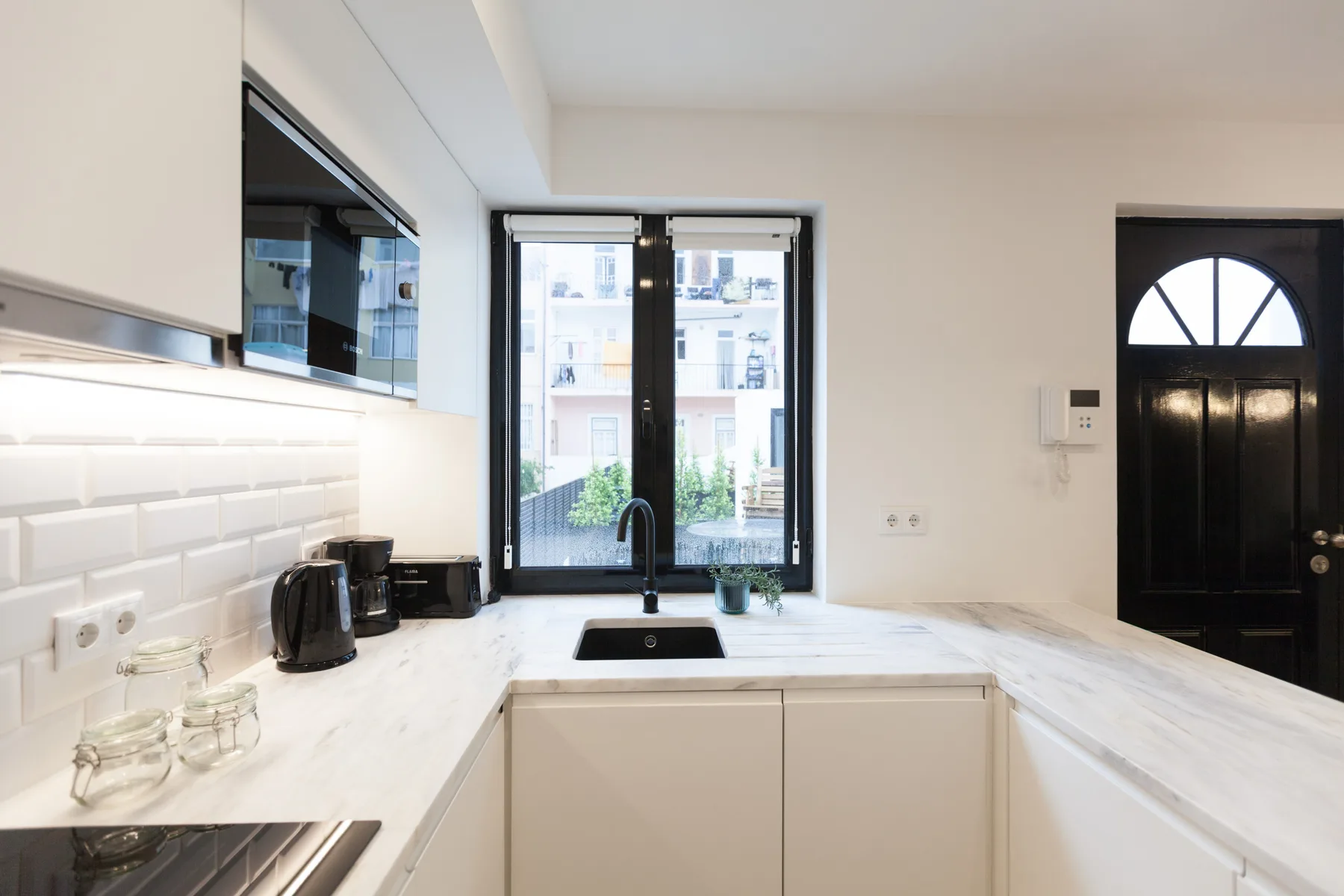 Una cocina luminosa con encimeras blancas, un fregadero y una ventana frente a edificios cercanos.