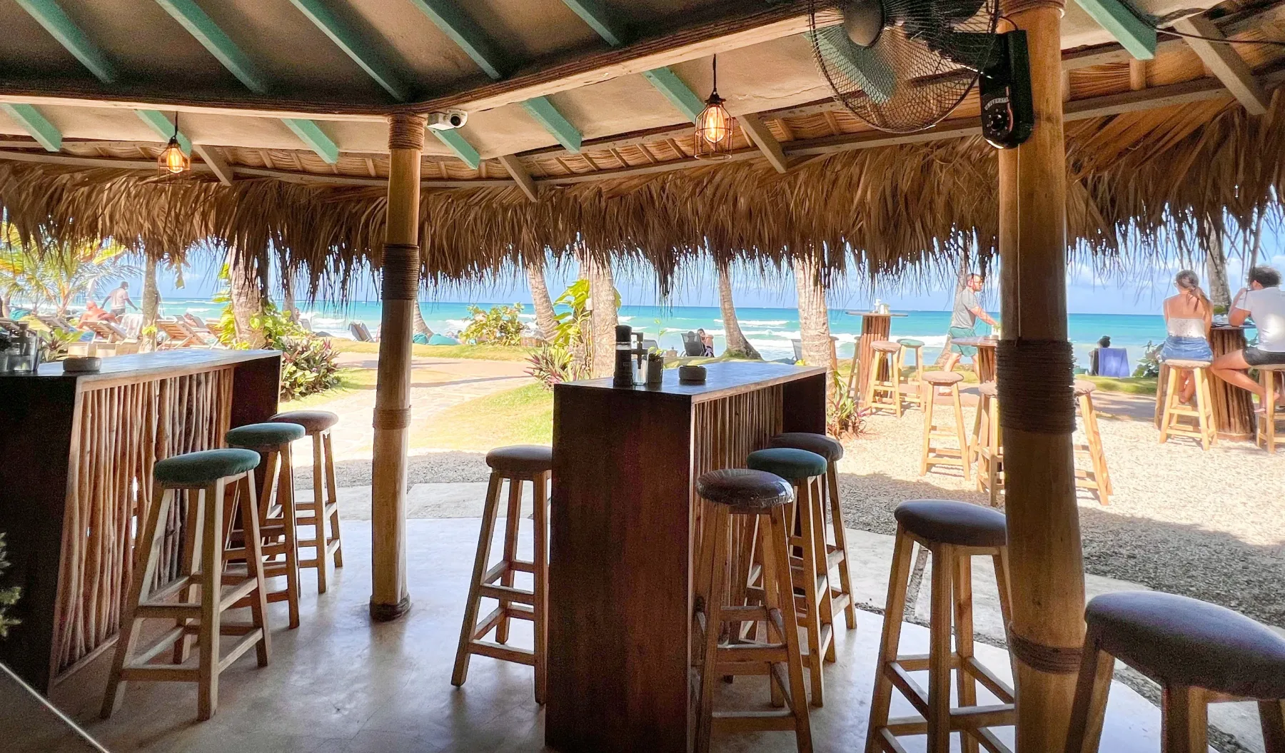Bar tiki frente a la playa con taburetes y vista al mar.
