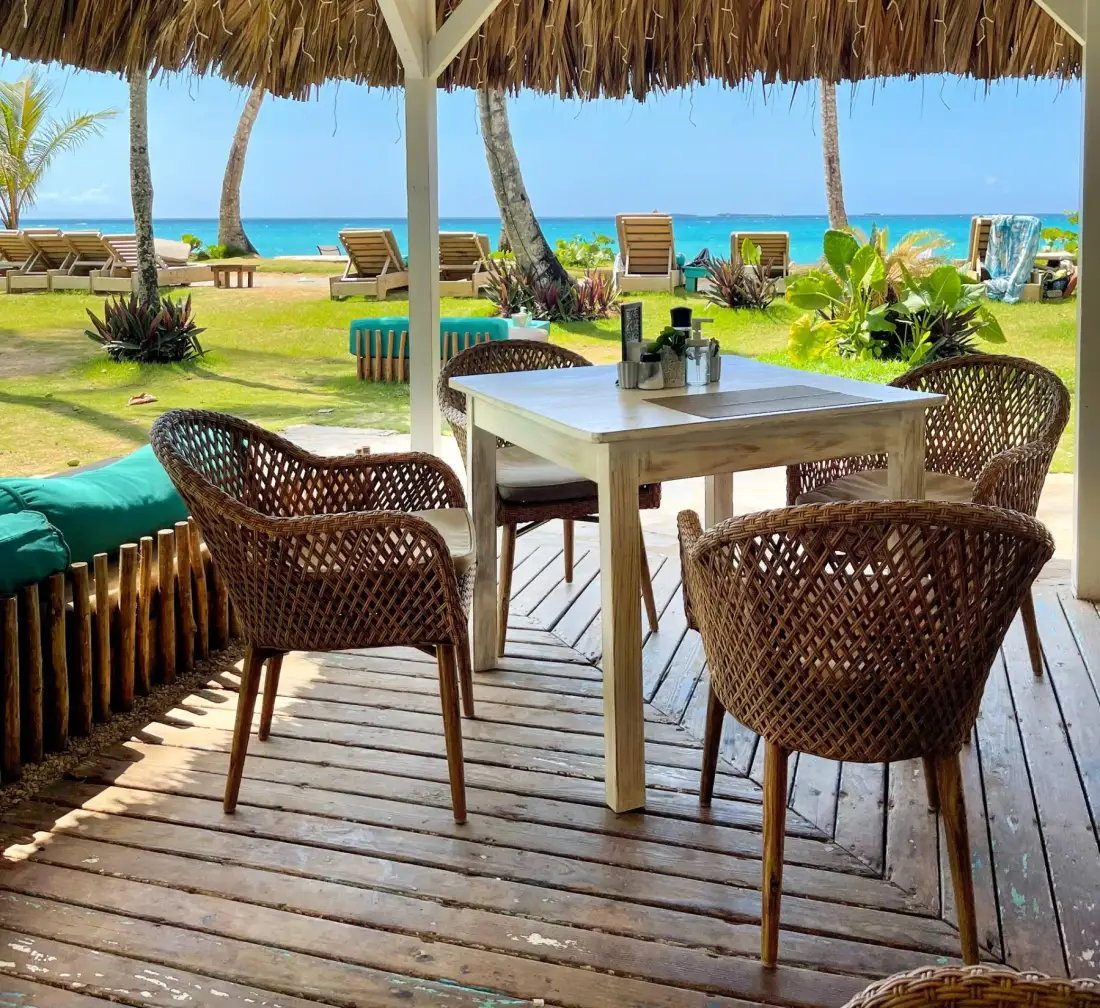 Beachfront seating area with wooden table and chairs under a thatched roof.