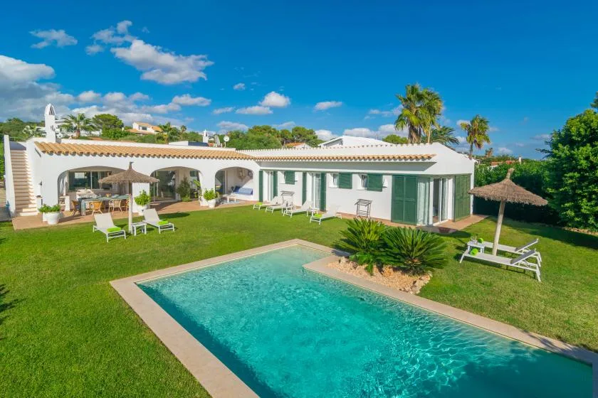 Una villa blanca da a una piscina y un césped bajo un cielo azul despejado.