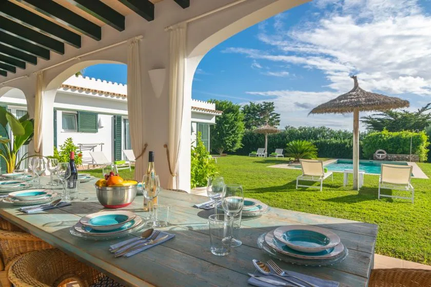 Una mesa de comedor bajo un patio con arcos, junto a un jardín y una piscina.