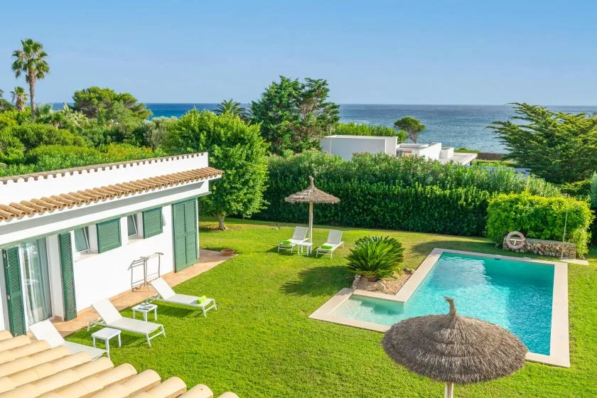 Un jardín rodea una piscina al aire libre junto a un edificio blanco, con el mar al fondo.