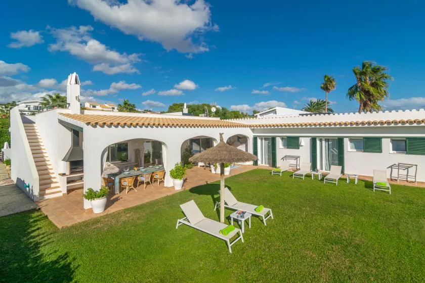Un edificio blanco rodea un patio con césped, asientos reclinables y una terraza sombreada.